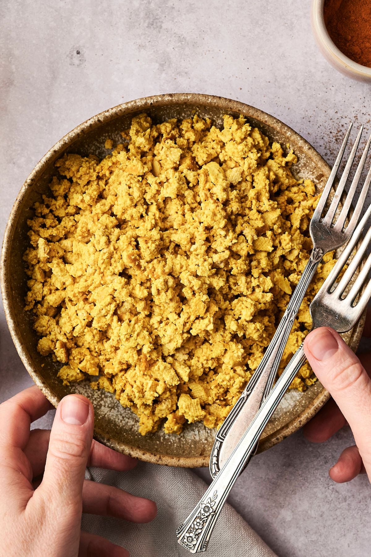 A bowl of scrambled tofu with a fork and spoon on the side. A hand is holding the bowl on a gray textured surface.