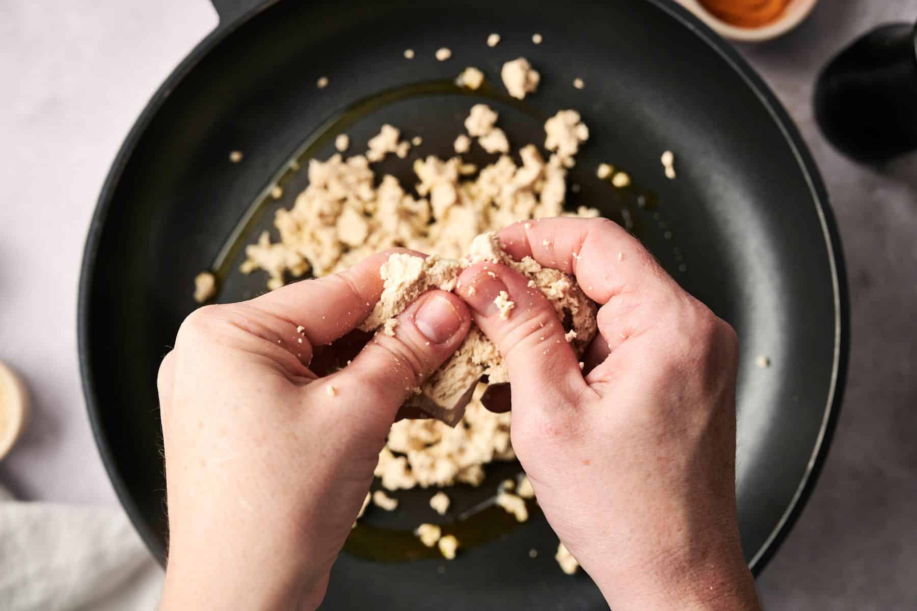 Hands crumbling tofu into a frying pan with oil.