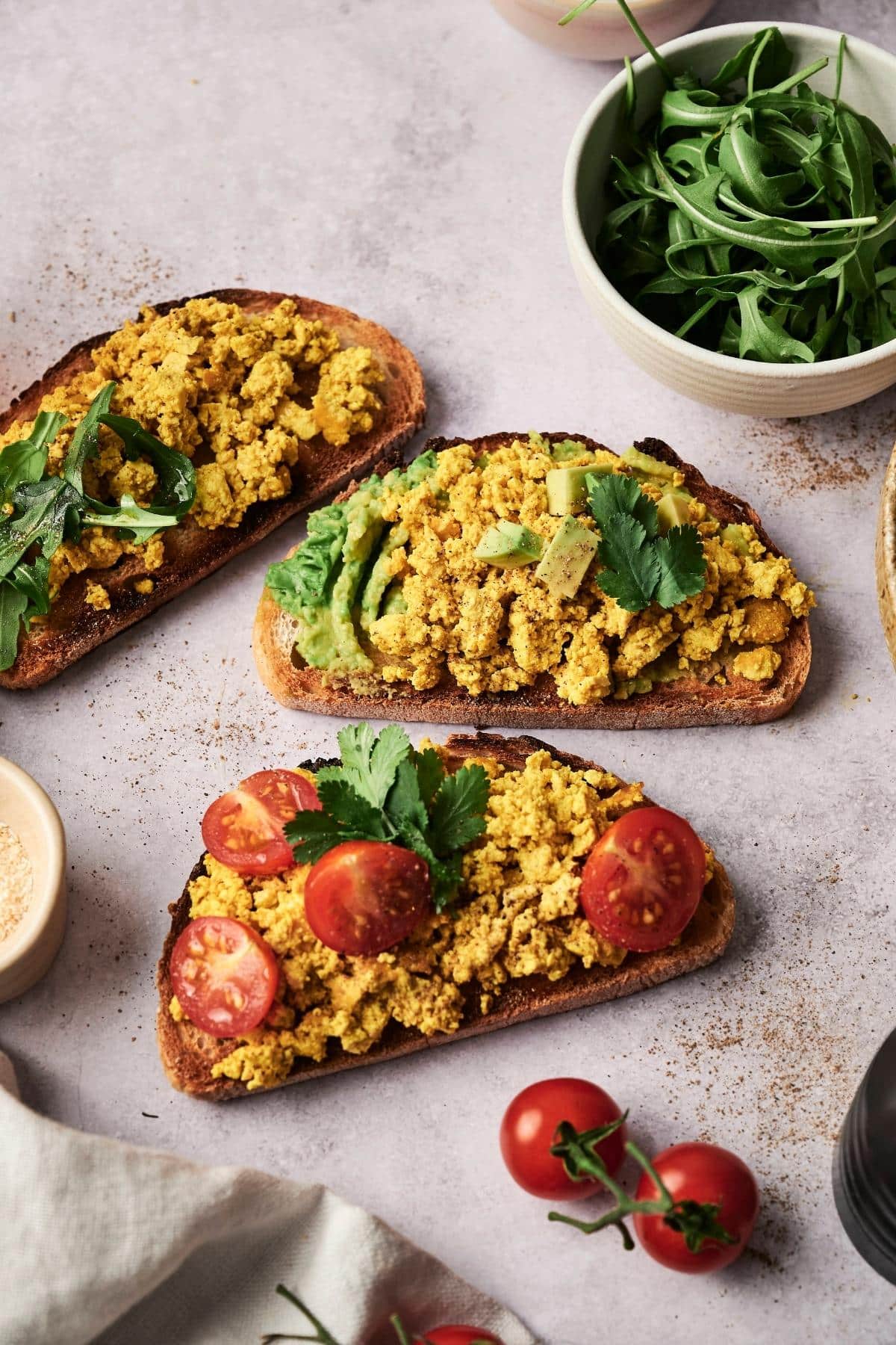 Three slices of toast topped with scrambled tofu, one with sliced avocado, one with cherry tomatoes, and one with greens, placed on a textured surface beside bowls of arugula and spices.