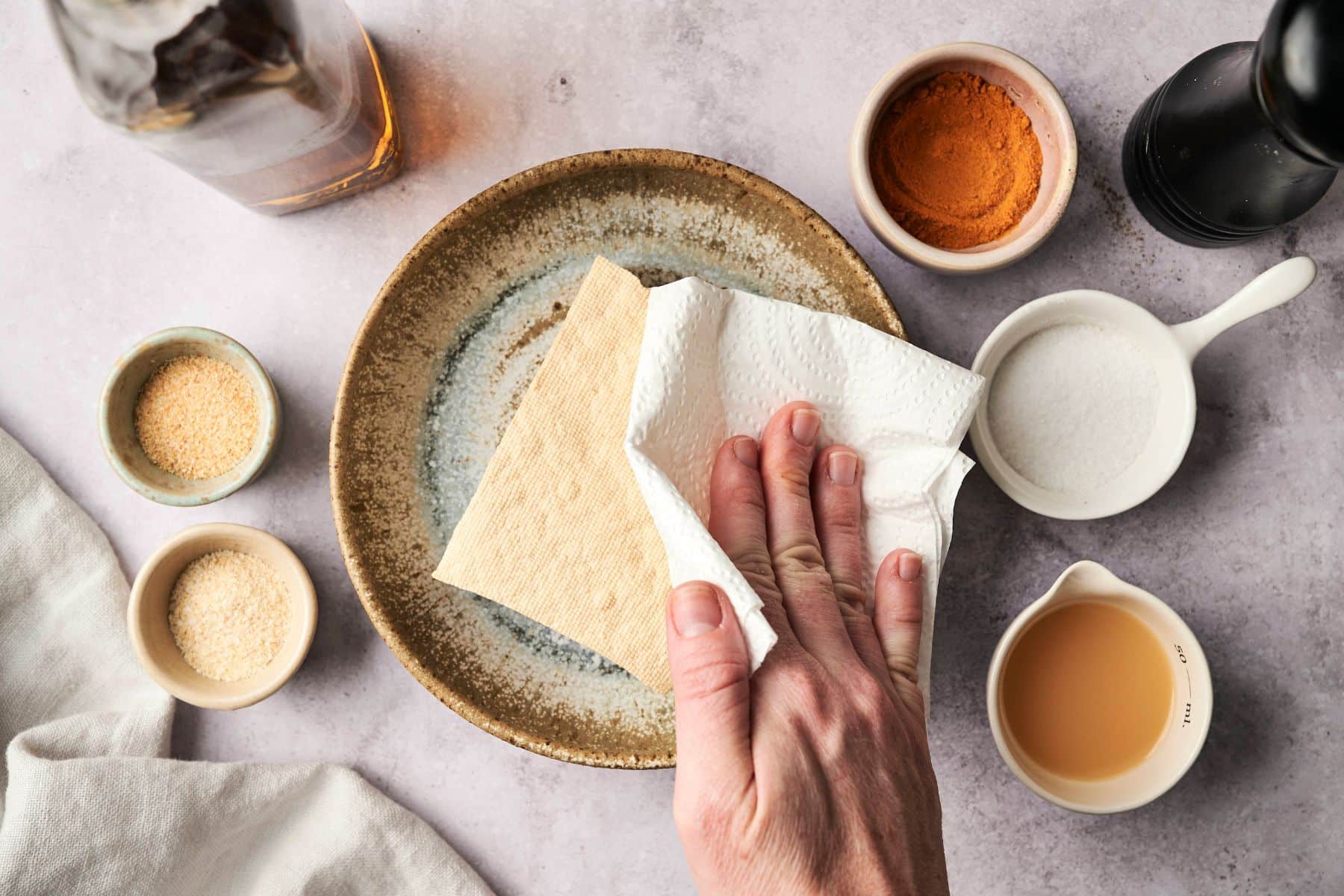A hand holding a paper towel pressing it into a block of tofu on a plate surrounded by small bowls of spices.