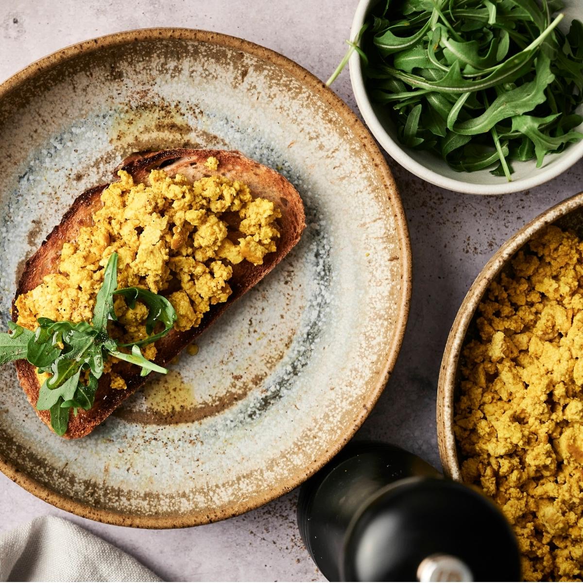 A plate with toast topped with scrambled tofu and arugula, a bowl of arugula, a bowl of extra scrambled tofu, and a pepper grinder on a textured surface.