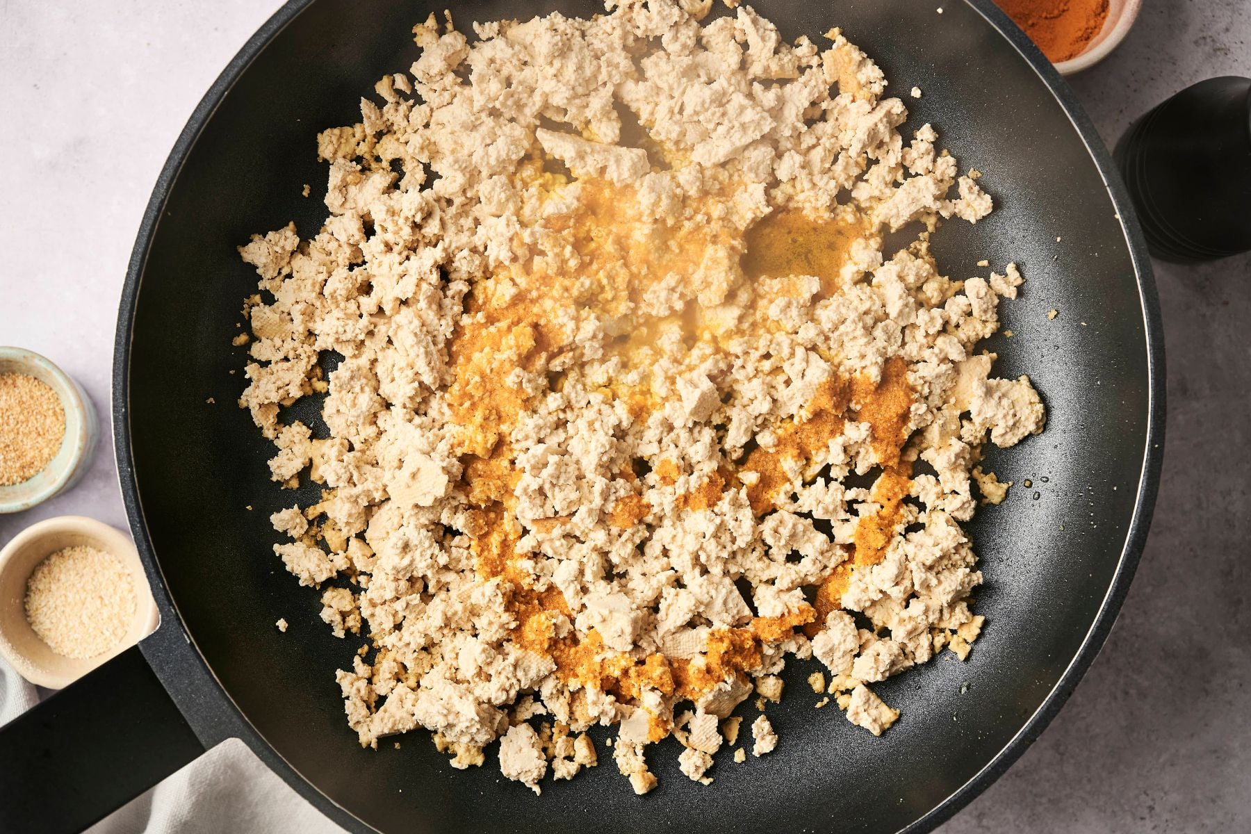 Crumbled tofu cooking in a black frying pan, with spices. Small bowls of sesame seeds and other seasonings are nearby on a light countertop.