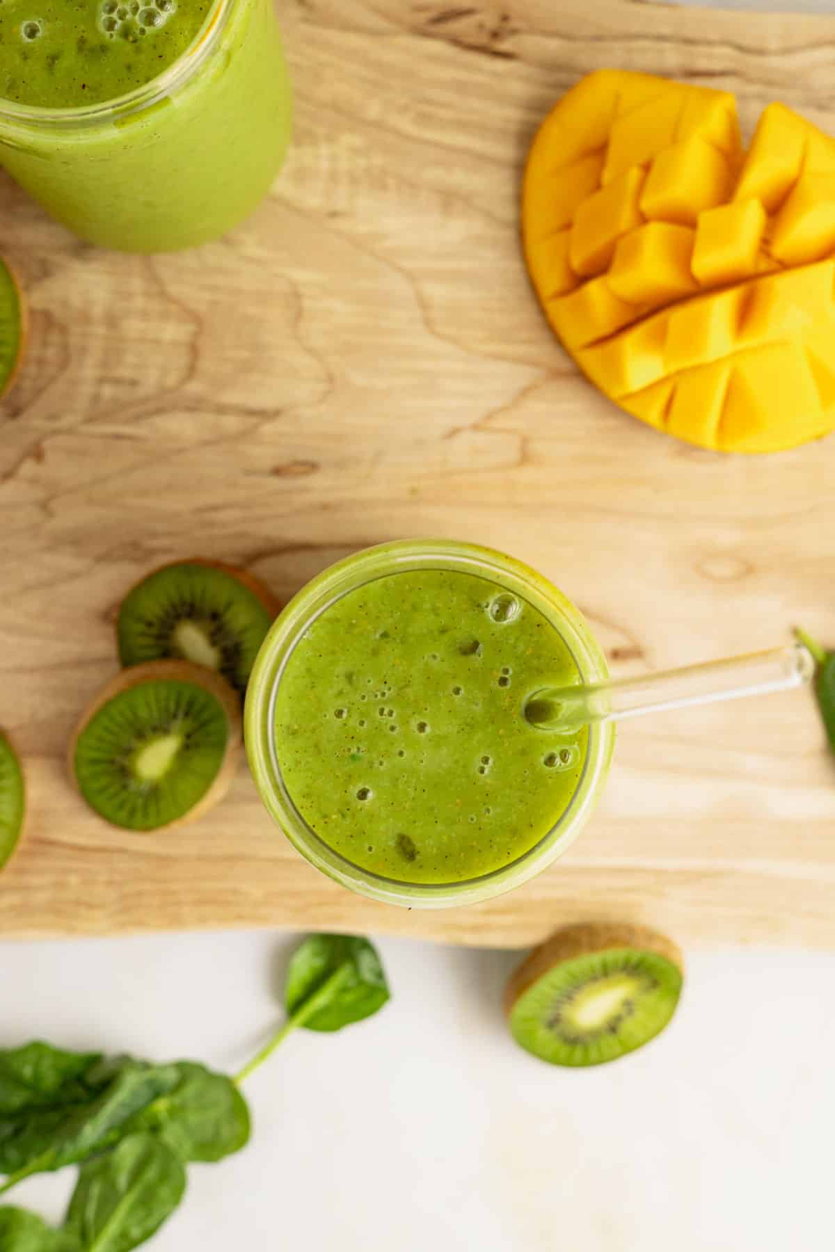 Green smoothie with fresh mango, kiwi, and spinach on top of marble and wooden cutting board.