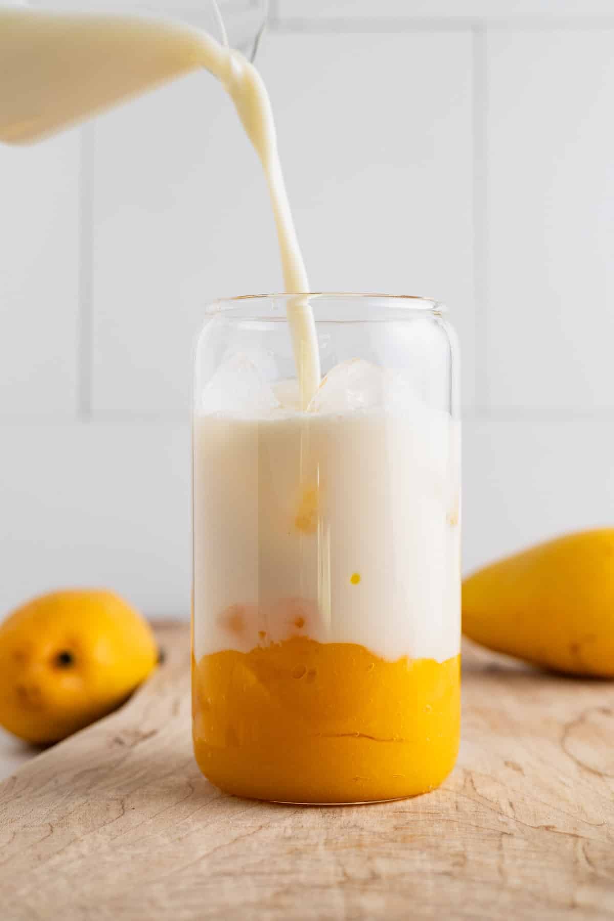 Milk being poured over ice in a clear glass cup with mango puree.
