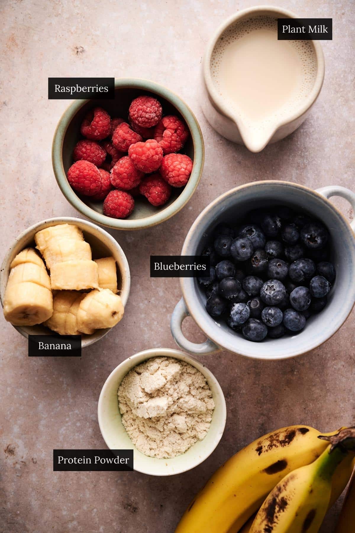 Bowls of berries, frozen bananas, protein powder, and almond milk all labeled.