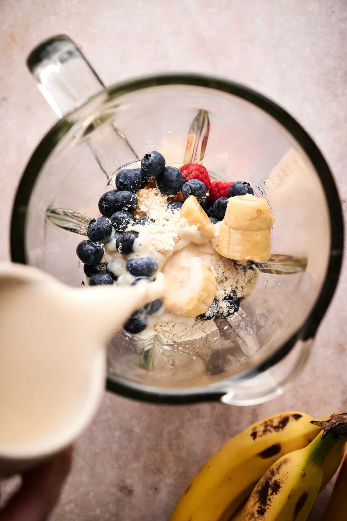 Milk being poured in a blender over top blueberries, raspberries, and bananas.