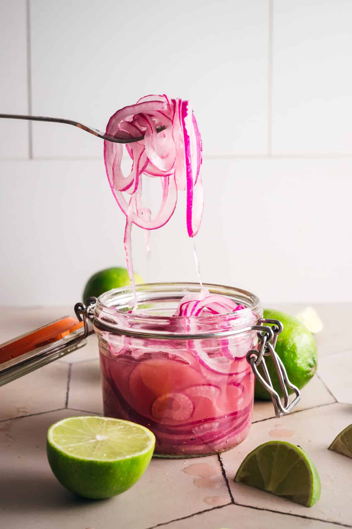 Pickled red onion with lime juice held on a fork in front of a tile backsplash.