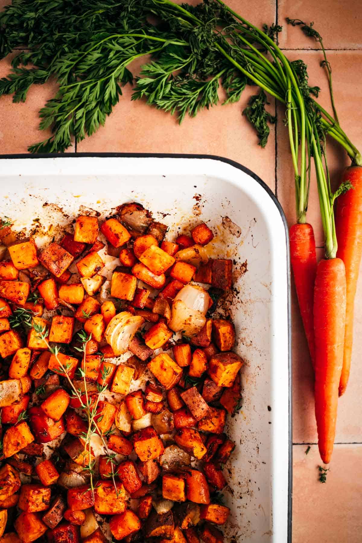 Baked root vegetables in an enamel pan next to fresh carrots.