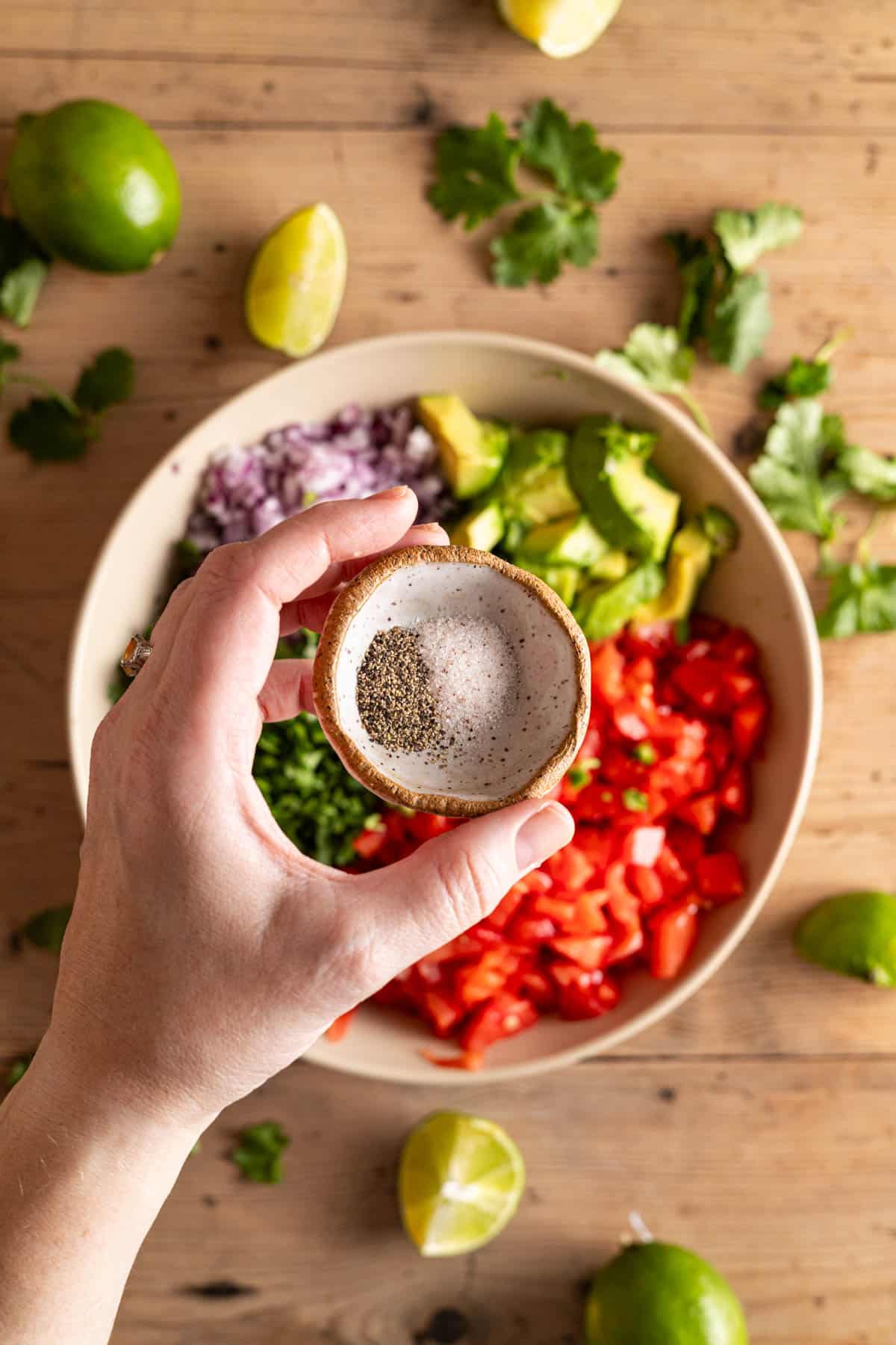Salt and pepper being squeezed into a bowl filled with chopped fresh veggies.