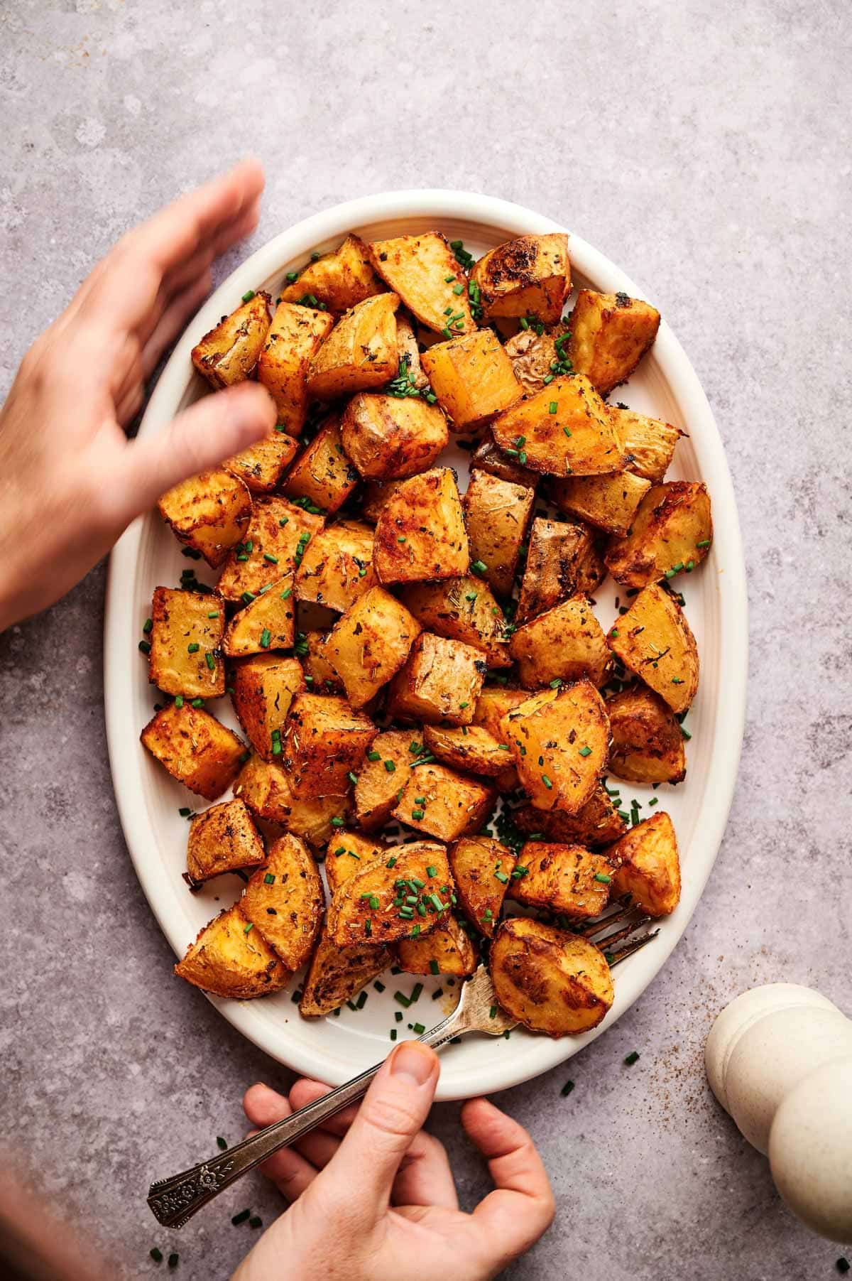 Hands reaching for oval plate of baked home fries.