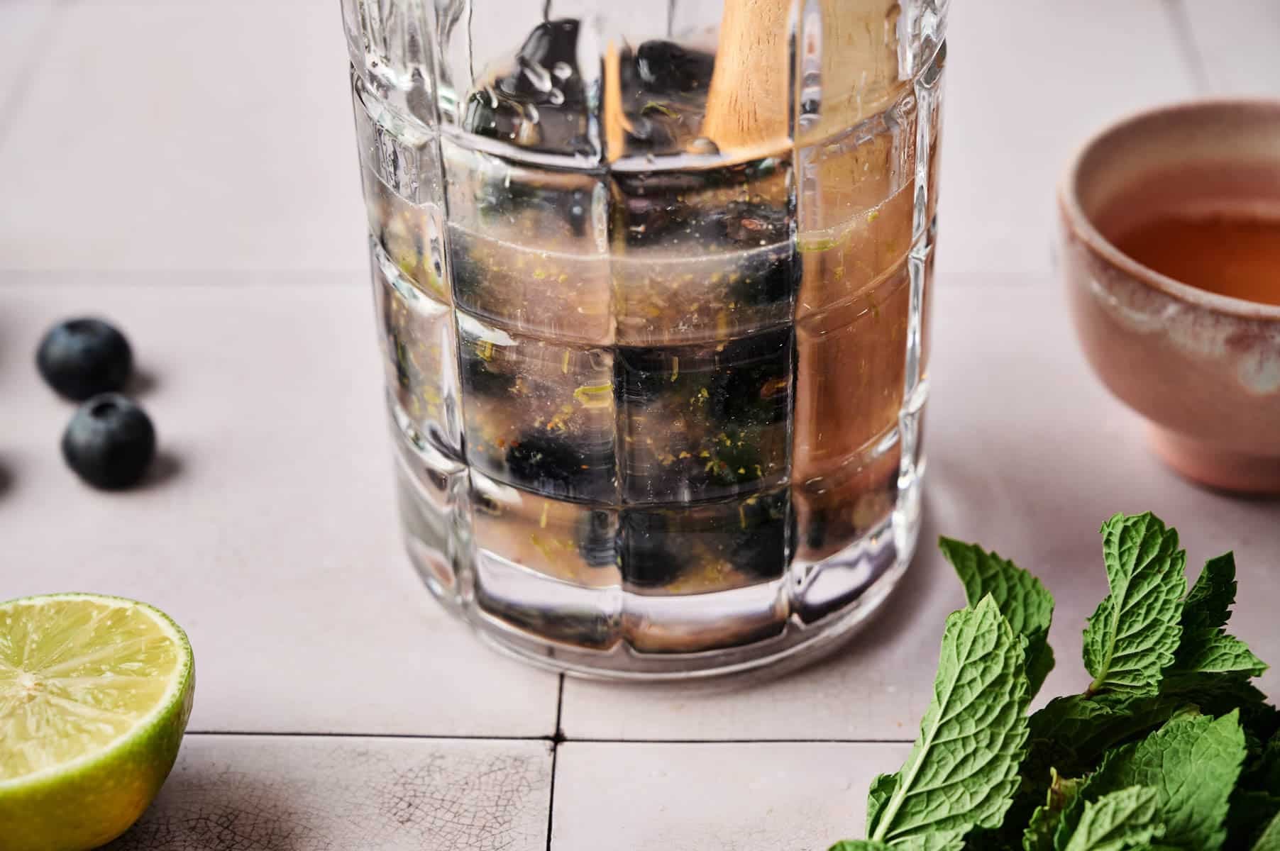 Mashing blueberries with a wooden muddle in a clear glass pitcher.