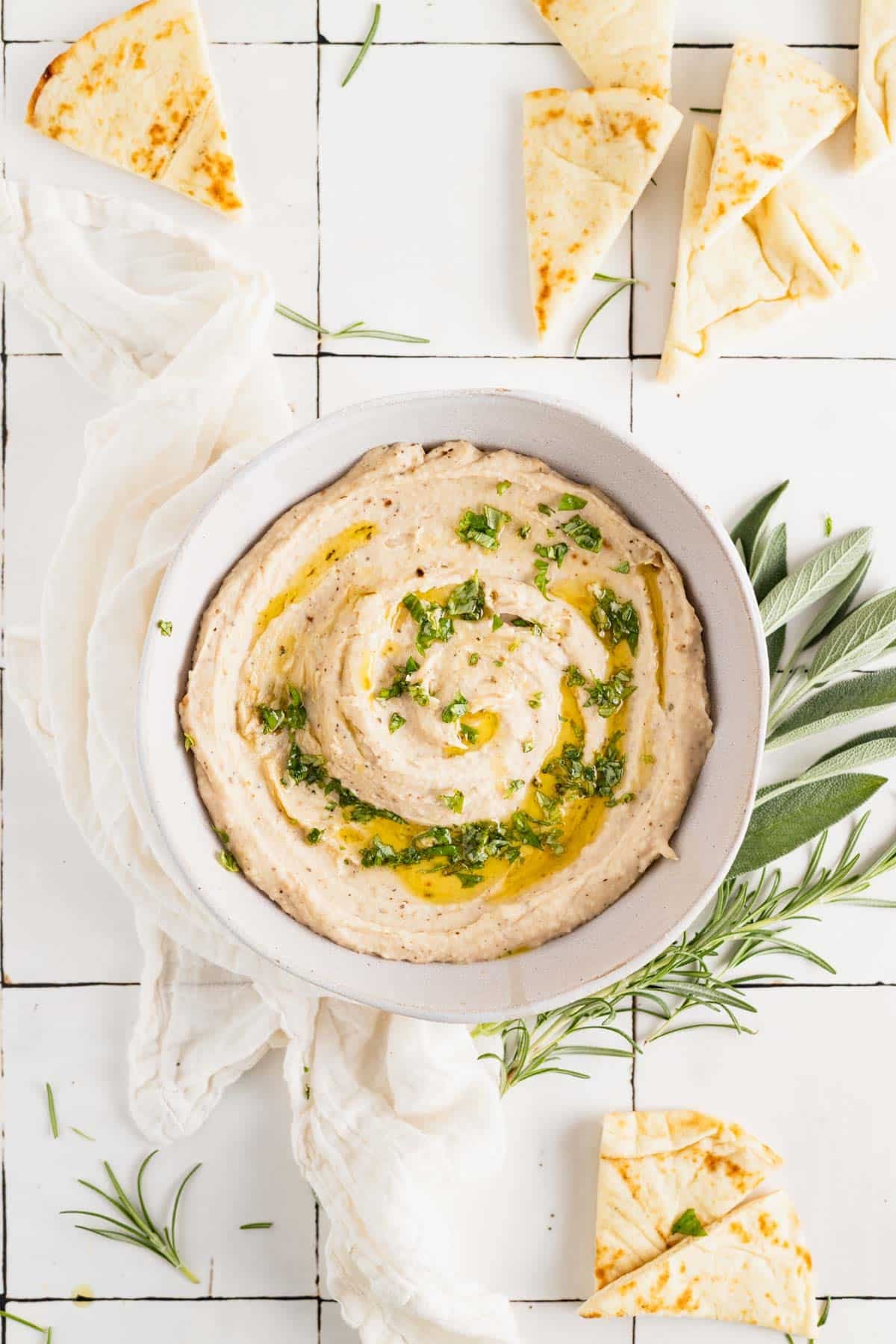 Homemade white bean dip in a white bowl with olive oil drizzled on top and fresh herbs.