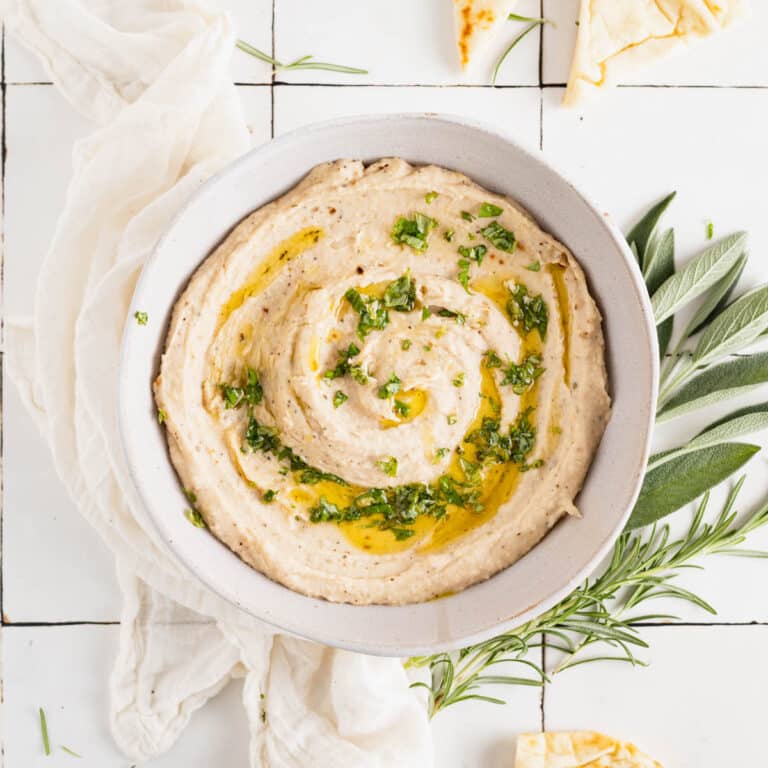 Homemade white bean dip in a white bowl with olive oil drizzled on top and fresh herbs.