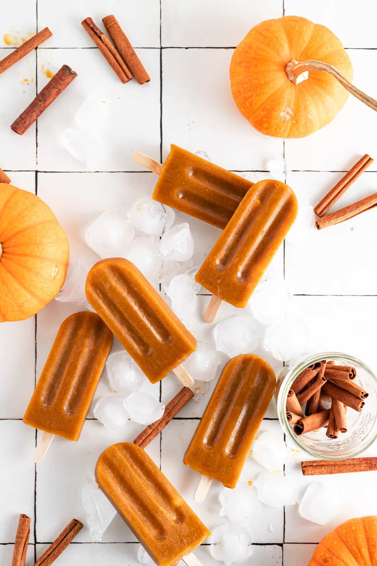 Pumpkin pie popsicles on a white tile surface with ice cubes and cinnamon sticks.